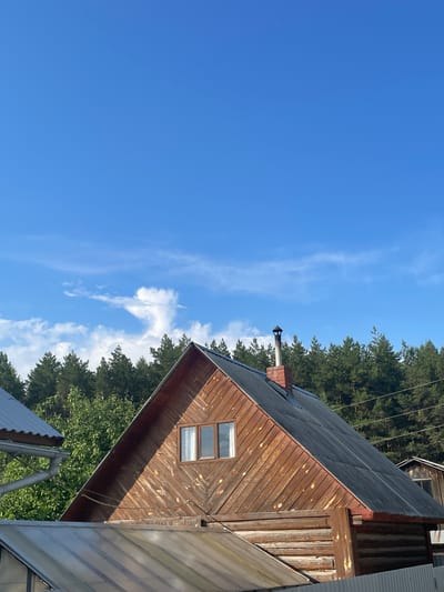 The image depicts a wooden house with a gabled roof in Zav'yalovo, Russia. The house appears to be constructed from logs, with a siding of vertical wooden planks on the upper facade. A chimney made of brick rises from the roof. Two windows are visible on the upper part of the house. In the foreground, other structures with corrugated roofs are partially visible. Behind the house, a dense line of evergreen trees forms the background, suggesting a rural or forested setting. The sky above is a clear blue with scattered white clouds. The lighting indicates it is daytime.