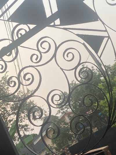 The image displays an upward-looking view through an ornate, dark metal gate or railing. The intricate metalwork, likely wrought iron, is predominantly composed of decorative spirals and scrolls of various sizes, with some larger, more angular and geometric shapes visible on the left side of the frame. The metal is dark, creating a strong silhouette against the bright background.
Through the openings in the metalwork, a hazy, light grey or off-white sky dominates the background, suggesting an overcast day or a bright, diffused light. Scattered green foliage from trees and some broader leaves, possibly from banana plants, are visible beyond the gate, particularly towards the right side and lower parts of the frame. Faint outlines of residential structures, such as roofs and walls, can be discerned through the leaves and haze, mostly on the right. Thin, dark lines resembling power lines or utility wires also traverse the upper middle section of the background.
The lighting creates a high-contrast scene, with the dark, silhouetted metalwork in sharp focus, while the background elements appear softer and slightly blurred due to the bright light source being behind the gate. The overall impression is one of looking out or up from behind a decorative barrier into a hazy, daytime outdoor environment. There is no visible text in the image.