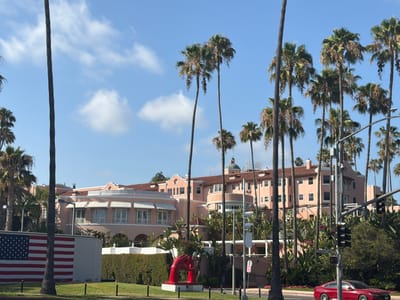 The image shows the Beverly Hills Hotel, a historic landmark located in Beverly Hills, California, United States of America. The hotel is a large, pink, Spanish Colonial Revival style building with red tile roofs, surrounded by lush greenery and numerous palm trees. In the foreground, an American flag is displayed on a wall. A bright red sculpture of two hands reaching for each other is also visible in front of the hotel. The sky is blue with a few white clouds. The scene appears to be a sunny day.