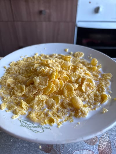 A close-up, overhead shot shows a white plate filled with corn flakes and milk. The plate has a faint green botanical design near the bottom left. The breakfast is set on a patterned tablecloth. In the background, a brown wooden cabinet is visible, along with the edge of a white oven with a black door. The image appears to be taken indoors, likely in a kitchen, possibly in a private residence in the city of Vetluzhsky, Russia, given the presence of common household items and Russian cabinetry. There are no people or animals in the frame.