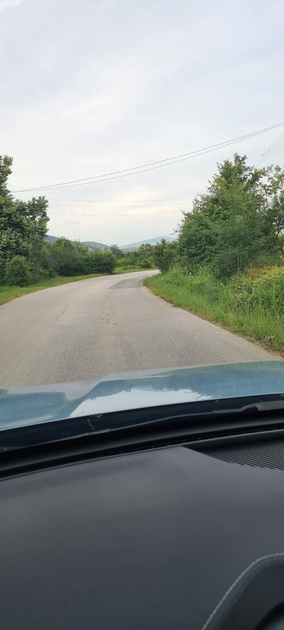 The image shows a view from inside a vehicle, looking out at a paved road winding through a lush green landscape. The road curves to the left and then to the right, disappearing into the distance. On either side of the road are dense trees and vegetation, with rolling hills and mountains visible in the background under a cloudy sky. Power lines are strung across the sky. The front of the vehicle, including the dashboard and windshield, is visible in the foreground. The scene appears to be in a rural area, possibly outside of the city of Laga, Bulgaria. There are no people or animals visible in the image. The overall impression is one of a scenic drive through the countryside.