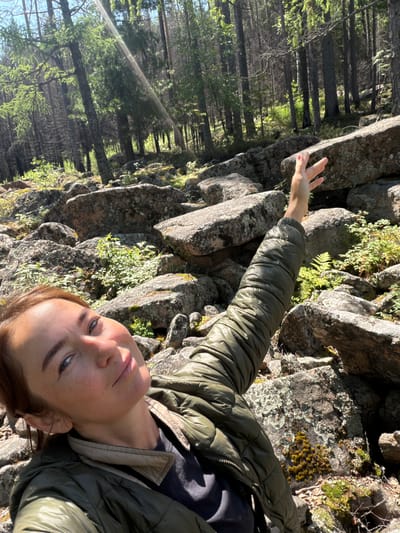 A woman with auburn hair and freckles, wearing a green puffer jacket, is taking a selfie in a forest. Her left arm is raised, with her hand open and extended towards the sun, which creates a light flare across the image. The background consists of a rocky terrain with trees and greenery. The location is identified as Orlenok, Russia.