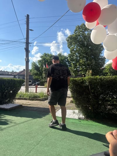 The image depicts an outdoor scene on a clear, sunny day in Targovishte, Bulgaria. A male subject, viewed from behind, is the central figure, walking towards the street. He wears a black short-sleeved t-shirt with a white graphic design on the back, which includes the text "HUGO" and stylized patterns. He also wears dark grey denim shorts, black sneakers, and white ankle socks.
In the foreground, a green, textured, artificial turf-like surface covers the ground, with scattered white decorative stones bordering it to the left and right. A long shadow, presumably cast by the male subject, stretches across this surface. A hand is partially visible in the bottom right corner of the frame.
The midground features a paved sidewalk and a street. A prominent concrete utility pole stands on the left side of the street, laden with multiple black electrical wires and junction boxes; a street lamp is affixed to it. A red fire hydrant is visible on the sidewalk near the base of the pole. Across the street, residential buildings with tiled roofs and varied facades are visible, along with parked vehicles, including a white van and a dark car. Green hedges and various bushes line the street and properties.
The background consists of numerous deciduous trees with dense green foliage, set against a clear blue sky dotted with scattered white cumulus clouds. In the upper right quadrant, a cluster of inflated balloons, predominantly white with several red ones interspersed, floats in the air, partially obscuring the trees and sky. Faintly visible in the distant background, to the right of the utility pole, are several flags on poles, one of which appears to be red and white.