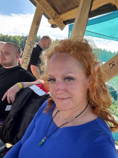 This image captures an outdoor scene under a partial wooden shelter in Большая Кивара, Russia, on a sunny day. In the foreground, a woman with voluminous, curly red hair and distinct pointed prosthetic ears (elf-like) is visible. She wears a long-sleeved blue top and a black cord necklace with a green, faceted pendant. Her eyes are light-colored, and she has visible eyelashes. She is looking directly at the camera with a subtle smile.
To her left, a man with a shaved head and beard is partially visible, wearing a black t-shirt. He has a yellow wristband and a black wristband with white text (identifiable as "ЯННИКА" in Cyrillic script) on his left arm. His right arm rests on a black jacket or bag, and he is looking away from the camera to his left.
Further in the background, another man is partially obscured by a wooden beam and the foreground woman's hair. He has a beard with a green substance on it, possibly paint, and is wearing a black shirt, looking downwards.
The scene features prominent wooden beams of a structure. One vertical beam has the Cyrillic text "ТАНЯ М" written vertically in black. An overhead horizontal beam displays three hand-drawn heart shapes. A turquoise-colored tarp or canopy is visible in the upper right, providing shade. The background shows a clear blue sky with white clouds and a dense line of green forest in the distance, suggesting a natural, open environment.