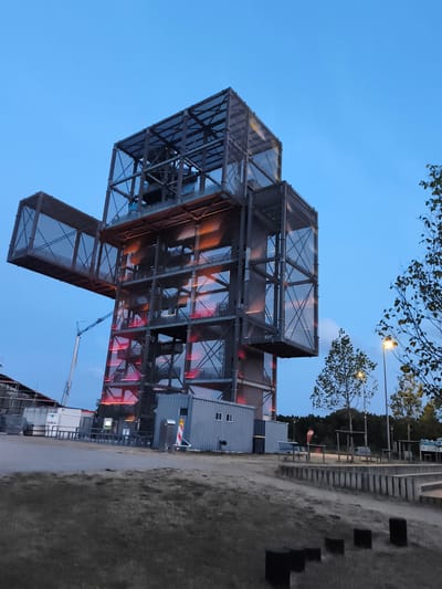 The image depicts the Indemann, a public art installation and observation tower located in Inden, Germany. The tower, designed in a cross-like shape, is constructed from a dark metal frame with illuminated orange and red lights visible within its structure. It has multiple levels and cantilevered sections. In the foreground, there is a grassy area with some dark, cube-shaped objects. To the left of the tower, a construction crane is partially visible, along with several white modular buildings and fencing. Further to the right, there are trees, a lamppost, and some outdoor seating. The sky is a deep blue, indicating it is either dawn or dusk. The scene appears to be an outdoor recreational or exhibition space.
