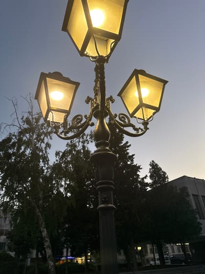 The image shows a multi-headed lamppost with glowing yellow lamps against a dusky sky. The lamppost is ornate, with decorative metalwork. In the background, silhouetted trees and a building are visible. The time of day appears to be dusk or early evening, as the sky is a pale blue transitioning to a lighter hue near the horizon. The building in the background has several windows, some of which appear to be lit. The overall scene is an outdoor urban or park setting. There is no discernible text in the image. The image evokes a sense of twilight calm.