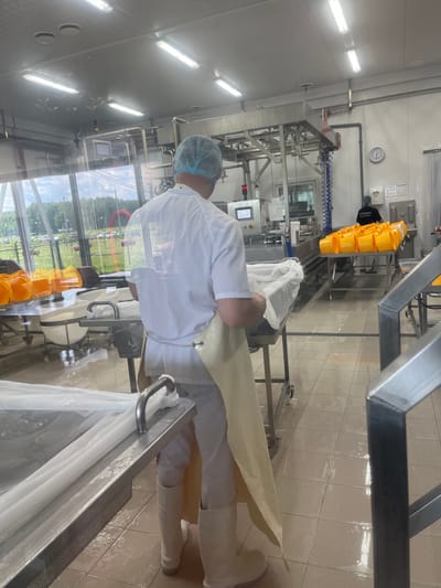 The image shows a man wearing a white uniform, a blue hairnet, and a yellow apron in a food processing facility. He is standing in front of a stainless steel table covered with white plastic sheeting, and appears to be working with it. The room has a tiled floor, industrial equipment, and large windows offering a view of a green outdoor area with trees and parked cars. Another person in a dark shirt with text is visible in the background, near a table laden with yellow buckets. A clock on the wall indicates it is approximately 11:44. The setting is consistent with a food production or preparation area in Akhlebaikha (Kovriginskii S/S), Russia.