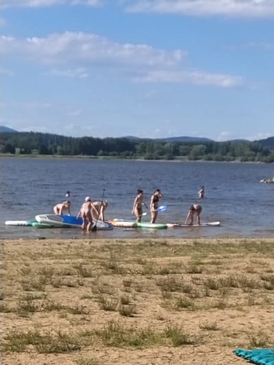 In Radslav, Czechia, on a clear, sunny day, several people are enjoying a lake with paddleboards. The foreground shows a sandy area with dry grass. In the water, multiple individuals are interacting with paddleboards. On the left, two people are standing on and near a blue and white paddleboard, with one person appearing to be reaching for something. To their right, three people are clustered around a blue and white paddleboard, with one person bending over, and another appearing to be adjusting something. Further to the right, two people are on separate paddleboards, and another person is standing in the water in the distance. The lake is calm, and a line of trees with mountains in the background borders the far side of the water.
