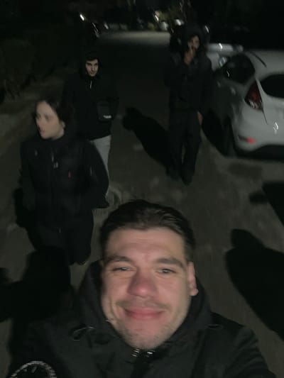 The image displays four individuals in an outdoor, dimly lit, nighttime setting within the Gerakas Municipal Unit, Greece. In the immediate foreground, a male subject with dark hair, a mustache, and a slight beard smiles faintly while looking directly at the camera, wearing a dark hooded jacket with a visible emblem on the left sleeve. Behind him, from left to right, are three additional subjects. A female subject, with dark hair, is positioned to the left, looking towards her left, dressed in a dark puffer vest over a dark long-sleeved garment. Centrally located behind the foreground subject is a male subject wearing a dark hooded jacket, light-colored pants, and a dark cross-body pouch, looking towards the camera. To the right, a fourth male subject, in dark attire, is partially visible and appears to be walking away from the camera. The subjects' elongated shadows are cast on the dark paved surface, originating from a light source near the camera. A white sedan is parked on the right side of the street in the mid-background. Distant streetlights or vehicle lights are blurred in the background.