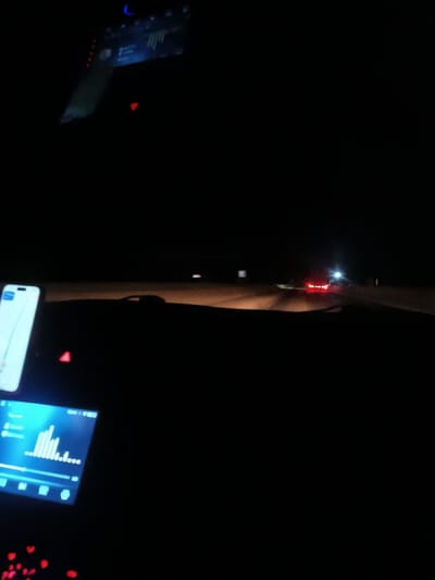 The photograph depicts a nighttime scene from inside a vehicle. Illuminated by the headlights, a road stretches into the darkness, with faint outlines of roadside signs visible. Taillights of another vehicle appear in the distance. Inside the car, a smartphone displaying a navigation app is mounted on the dashboard. Additionally, a dashboard screen glows with equalizer bars. The overall tone is dark and suggestive of a road trip at night.