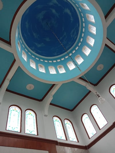 The image shows the interior of a mosque, specifically the dome and a portion of the walls. The dome is painted a bright blue, mimicking the sky with white clouds, and contains numerous stained-glass windows. A light fixture hangs from a single wire extending from the dome. The walls are white with dark brown trim around windows. There are no people or objects in the image. The location is Kecamatan Brangsong, Indonesia, based on coordinates (-6.948, 110.231).