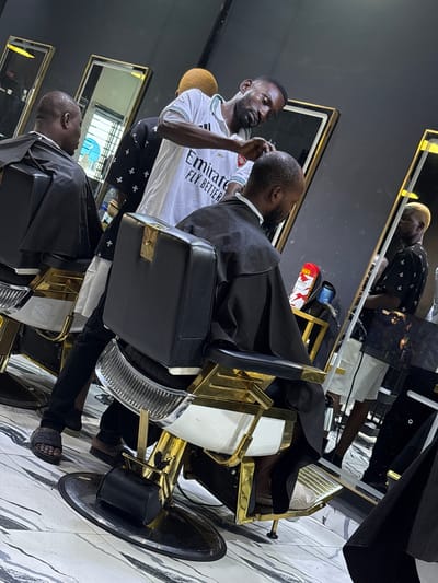 A man in a white Emirates t-shirt is giving a haircut to a seated man in a black barber's cape in a barbershop. The barbershop features multiple mirrors, gold-accented barber chairs, and a black ceiling. The location is Lugbe, Nigeria.