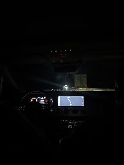 The image depicts the interior of a vehicle at night, viewed from a rear passenger perspective, looking forward through the windshield. The location is Rashkovo, Bulgaria.
The dark silhouette of a driver's left shoulder and arm is visible on the left, with hands gripping the steering wheel. The dashboard features an illuminated instrument cluster displaying red-accented dials, including a speedometer and tachometer. A central infotainment screen, exhibiting a prominent diagonal crack across its surface, displays a navigation map. Below the screen are climate controls and other vehicle function buttons. An overhead console contains several illuminated buttons, notably a red "SOS" emergency call button. The dark silhouette of a front passenger's right shoulder is partially visible on the right.
Through the windshield, a dark road at night is illuminated by the vehicle's headlights. Directly ahead is the rear of a light-colored commercial semi-trailer truck, bearing the inscription "BG Fardatrans" on its rear doors, with multiple red taillights active. Distant headlights from another vehicle are visible further down the road beyond the truck. The scene indicates the vehicle is stationary or moving slowly behind other traffic on a road at night.
