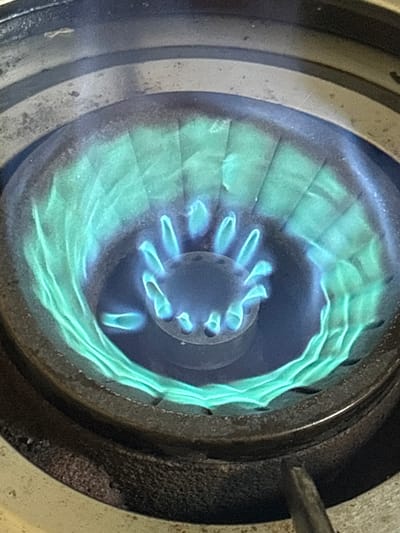 This is a close-up, high-angle shot of a gas stove burner with blue and green flames. The burner itself appears to be made of dark metal with some visible wear and grime. The flames are emanating from holes in the center of the burner, forming a circular pattern. The outer edge of the burner has a textured, metallic finish. The background is dark and out of focus, but appears to be part of a stove top. The image does not provide any information about the city of Lugbe, Nigeria, or any people, actions, or interactions.