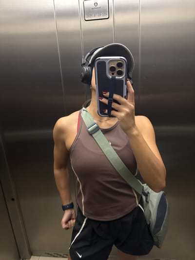 A woman is taking a selfie in an elevator. She is wearing a brown tank top, black athletic shorts with a white stripe and a Nike logo, headphones, and a smartwatch. She has a bag slung across her body and is holding a smartphone, using it to take the picture. The elevator has metallic walls, and a panel with some text is visible on the left side. The lighting is artificial and casts a slight shadow. The image is oriented vertically, indicating the woman is likely standing. The overall mood appears casual, perhaps after a workout. There is no visible indication of the time of day or weather outside.
Visible text on the panel reads: "420 KG e BEVG".
