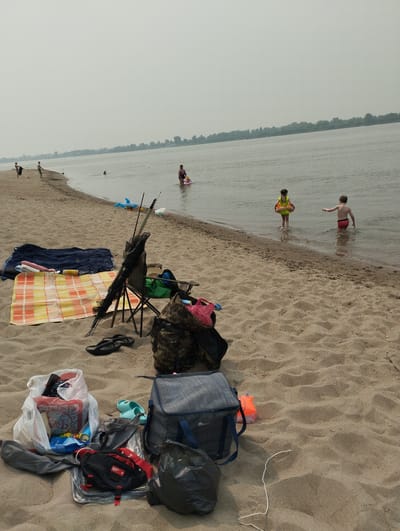 The image depicts a sandy beach with a body of water in the background. Several individuals are present in the water, including two children, one wearing a yellow life vest and the other in red shorts. An adult woman in a purple swimsuit is also in the water, near a pink flotation device. On the beach, a variety of items are laid out, suggesting a picnic or outing. These include beach towels, a blue net, a folding chair with fishing rods, a camouflage bag, a blue cooler, several plastic bags containing items, a black trash bag, and a pair of flip-flops. The sky is overcast, and a tree line is visible in the distance across the water. The scene appears to be a recreational area, likely a beach on the Ob River in Bibikha, Russia, given the context provided.
