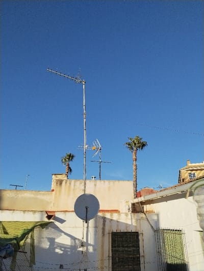 A daytime view of rooftops with antennas against a clear blue sky. A large antenna dominates the scene. Two palm trees are visible behind the roofs, adding a tropical touch. One satellite dish is attached to a wall. Buildings have windows with bars, and green shading on some parts. Overall, a blend of urban and tropical elements.