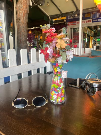 The image displays a detailed view of a dark wooden table in an outdoor or semi-outdoor cafe setting located in Kartal, Turkey.

On the table, from left to right:
A pair of round-framed sunglasses with dark lenses and gold-colored frames rests horizontally. The brand "PRADA" is visible, inverted, on the left lens.
To the right of the sunglasses, a clear glass vase contains a collection of multi-colored, irregularly shaped decorative stones or candies at its base. Artificial flowers in shades of red, pink, orange, white, and green, along with green leaves and small red berry-like accents, are arranged within the vase.
Behind the vase, a cylindrical silver-colored metal holder contains several white paper packets, likely sugar or condiments.
To the right of the holder, a dark-colored smartphone lies face down on the table.
Further to the right, a second pair of sunglasses, possibly aviator-style, with dark lenses, rests on a circular, lidded, silver-colored metal ashtray. The brand "Ray-Ban" is visible on one of the lenses. A light-colored strap, likely from a bag, is partially visible behind the ashtray.

Behind the table, a white picket fence-style wooden divider separates the seating area from the background. To the right of the fence, a turquoise-colored upholstered bench or cushion is visible.

The background reveals an outdoor commercial area. On the far left, a tall tree with reddish-brown bark stands next to a glass-fronted display refrigerator containing various bottled beverages. Behind the fence and across from the table, a commercial establishment with an awning features multiple signs. One partially obscured sign reads "KAPATLANDIR". A prominent blue sign displays the Red Bull logo. Below this, another sign clearly reads "MEKEZ BÜFE Tekel Bay". These signs identify the location as a buffet/kiosk and a tobacco/alcohol dealer in Turkey. Decorative lighting fixtures are suspended from the awning, and various artificial plants, including purple flowering ones, contribute to the cafe's ambiance. No people are visible in the scene.