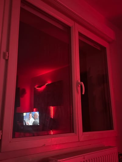 This image captures an interior residential or private room in Marcoussis, France, characterized by a dominant red ambient light. A white-framed, double-pane window occupies the central and upper-right portion of the frame. Reflected prominently on the lower-left window pane is a television screen displaying two adult males; one in a white lab coat observes a document or tablet held by a second male in a casual shirt. To the right of this reflection, a vertical array of red LED lights and a string of red fairy lights are visible, contributing to the room's red illumination. The window structure includes a white handle on the right pane and a white roller shutter housing above. A white radiator is positioned beneath the window. The precise location within Marcoussis, France, cannot be determined from visual cues within the image.