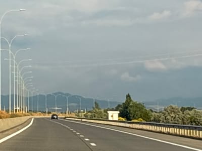 The image shows a highway in Samtsevrisi, Georgia, with a row of streetlights lining the left side of the road. A black car is visible in the distance on the highway. The road is paved and has white lane markings. To the right of the highway, there is a guardrail and some vegetation, including trees and bushes. In the background, there are mountains. The sky is overcast with some clouds.