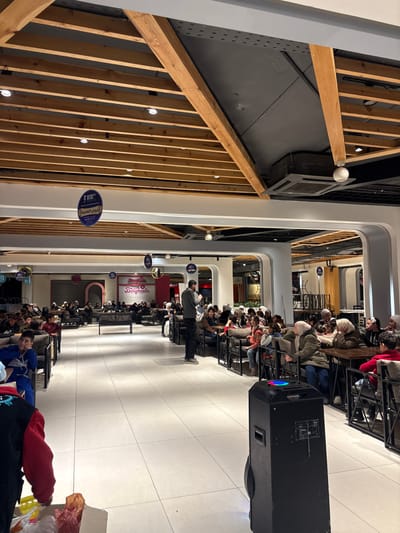 The image captures an indoor gathering or event, likely within a mall or large public building, characterized by contemporary architecture. The scene is viewed from an elevated perspective, looking down a long, brightly lit corridor or open space.
In the center of the frame, numerous people are seated in long rows facing towards the right side of the image (when oriented correctly). They are occupying chairs and stools positioned around what appear to be narrow tables or bar-style counters. Most of the attendees are looking forward, suggesting they are observing or listening to something or someone at the far end of the seating arrangement. One person, dressed in a light-colored top and dark pants, stands prominently in front of the seated audience, seemingly addressing them or presenting.
To the right of the seated audience, a large, black speaker unit stands on the tiled floor. It features colorful, illuminated lights on its side, indicating it is active and contributing to an event atmosphere.
The architectural design of the space is noticeable. On the left side, there's a prominent wooden slatted structure, forming a decorative wall or railing, with vertical planks and horizontal beams. Beyond this structure, a dark, possibly exposed ceiling or upper level is visible, with some ventilation ducts or industrial elements. The main pathway or floor where the event is held is covered with large, light-colored square tiles, reflecting the overhead lighting.
The space extends far into the background, revealing more rows of seating and additional architectural elements, including arched doorways or entrances to other sections. Several spherical light fixtures are suspended from the ceiling throughout the area, providing ample illumination.
Several circular blue signs with white text hang from the ceiling, one of which is clearly legible in the mid-ground. This sign displays both English and Arabic text: "7 days a week" and "7 أيام في الأسبوع" (7 days a week), along with "أصناف متنوعة" (various types). Further down the corridor, a red sign with white lettering is visible, displaying the word "Doughnuts" in a decorative font.
In the immediate foreground, partially cropped on the bottom, the back of a person wearing a red and blue garment is visible, along with a translucent plastic bag containing items, suggesting a casual presence in the periphery of the main event.
The overall impression is one of a well-attended public event or presentation taking place in a modern, multi-functional indoor venue.