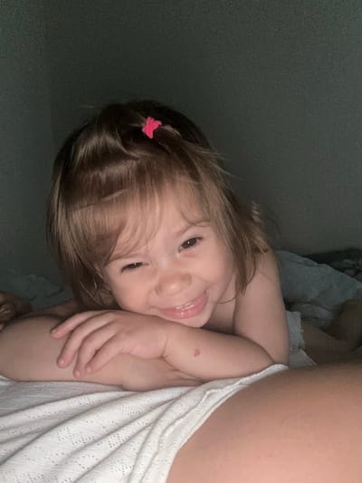 The media file is a close-up, slightly tilted, vertical photograph of a young child lying down. The child has brown hair, a small pink butterfly clip in their hair, and is looking towards the camera with a wide, happy smile, tongue slightly out. They are wearing a white knitted garment. The child's arm is resting on their cheek, and their other arm is visible. There is a reddish mark on their arm. The background is a dark, textured surface. The lighting suggests it might be indoors, possibly in the evening or a dimly lit room, given the overall shadow and the way the light falls on the child's face. The image conveys a sense of playfulness and happiness. No text is visible in the image. The location context provided is Kharnavy, Russia, but there are no specific visual cues within the image to confirm this location.