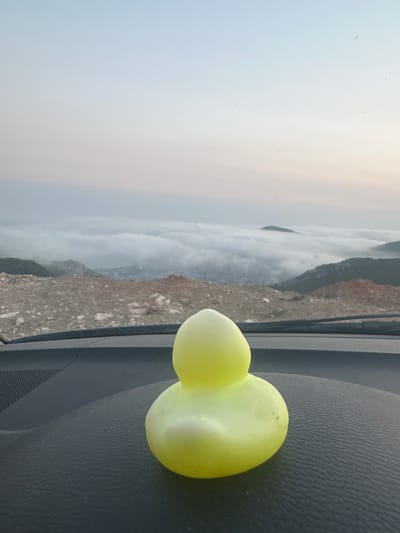 A yellow rubber duck toy is placed on the dashboard of a car. The car is parked on a rocky, barren hillside overlooking a valley. Below the hillside, a thick layer of clouds obscures the ground, with the tops of distant mountains visible above the clouds. The sky above is a pale blue, transitioning to a soft pink and white towards the horizon, indicating either sunrise or sunset. The overall scene suggests a quiet, elevated viewpoint, possibly in a rural or semi-developed area. The location is identified as Lapčići, Montenegro.