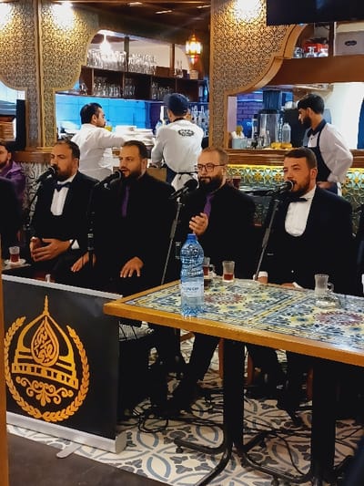 This image depicts an indoor scene within a restaurant or cafe setting in Dummar Municipality, Syria. In the foreground, four adult males are seated behind individual microphones, suggesting a performance or speaking event. They are formally dressed in black jackets; two wear black bow ties, and two wear purple ties or ascots. A wooden table with a multi-colored, patterned tile surface stands before them, holding a plastic bottle of "NAZANECHI" brand water and four small clear glasses containing a dark liquid. To the left of the table, a black banner displaying a gold emblem of a stylized dome with Arabic calligraphy and a laurel wreath is positioned on the floor. The floor beneath the table features light-colored geometric patterned tiles, with some visible wiring. In the background, three male staff members are visible behind a counter area; one in a white uniform is holding plates, another in a white shirt and blue cap is partially obscured, and a third in a white shirt and dark apron stands by a counter. The background also shows intricate, carved wooden decorative screens, shelves stocked with glassware, and warm-toned lighting from ceiling fixtures.