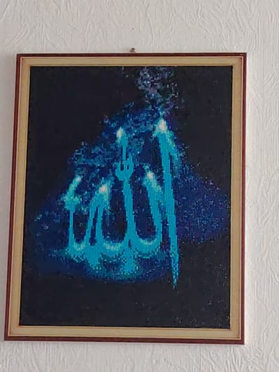 The image shows a framed artwork featuring blue calligraphy on a dark background. The calligraphy appears to be Arabic script, potentially religious in nature, with swirling patterns and light accents. The frame is a dark reddish-brown with a lighter inner border. The artwork is hung on a textured white wall. There are no people or other objects visible in the frame, and the background is indistinct. The location in Kogalym, Russia, cannot be determined from the visual information presented in the image.