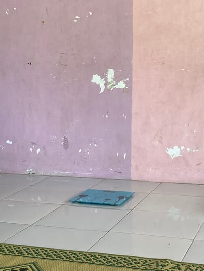 The image shows a bathroom scale, laying flat on a white tiled floor. The scale is blue and has black markings. The scale is situated in front of a wall that is painted in two shades of pink, separated by a vertical line. The wall has areas of peeling paint. The bottom of the image shows a portion of a green and brown rug. The location is a bathroom in Kecamatan Piyungan, Indonesia, at the coordinates provided.