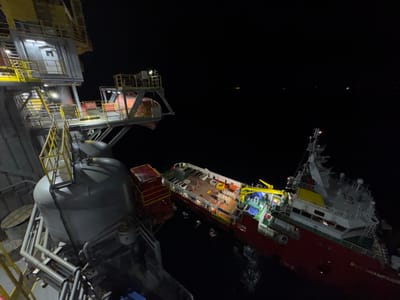 This nighttime aerial view captures an offshore oil rig or platform situated next to a red and white ship. The platform, with its yellow railings and industrial lighting, appears to be a large, complex structure. A significant part of the platform, including tanks and access ways, is visible in the foreground.
The ship, identified by the visible text "BUS CHAMPAGNE" on its hull, is docked alongside the platform. The ship is illuminated, revealing its deck with various equipment, cargo, and what appear to be a few people in orange high-visibility vests. A yellow crane is visible on the ship's deck. The overall scene suggests an operational environment at sea during the night. The water around the vessels shows some disturbance, indicating movement. The dark sky and the lights from the platform and ship are the primary light sources. The vast expanse of the dark ocean stretches out behind the two vessels, with distant lights suggesting other offshore installations or vessels. The image conveys a sense of industrial activity in a remote maritime setting at night.