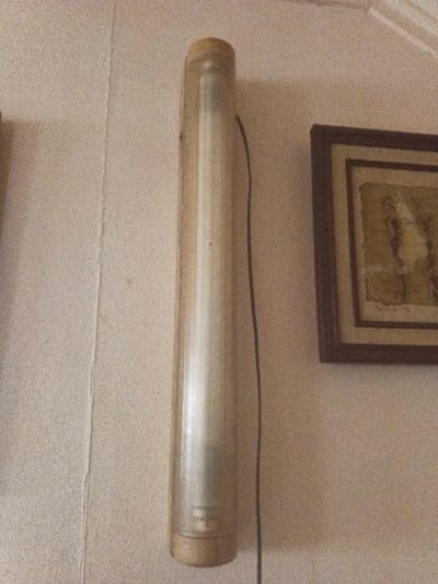 An interior shot depicts a vertical, cylindrical fluorescent light fixture with a translucent cover mounted on a light-colored, textured wall in Dogsenang Gorphoog, Bhutan. A black electrical cord runs parallel to the fixture. To the right, a dark brown framed artwork featuring abstract or textured elements in earthy tones is partially visible, also mounted on the wall. A vertical seam or crack is present on the wall to the left of the light fixture. No people are visible.