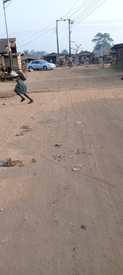 A sandy road dominates the foreground, marked with tire tracks and scattered debris. A person hurries across the road carrying a load on their head. In the background, a silver car is parked beside weathered shacks with corrugated roofs. People and motorcycles gather near the shacks. Power lines stretch across the sky above. The scene depicts a rural or impoverished setting with basic infrastructure.