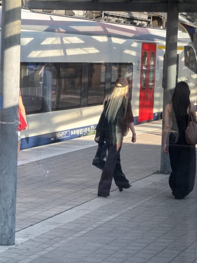 The image shows a train station platform in Leuven, Belgium, with a train stopped alongside. Two women are walking on the platform. The woman on the left has long blonde hair, is wearing a dark top, wide-leg dark pants, and a brown cap. She is carrying a dark bag. The woman on the right has long dark hair and is wearing a dark dress and carrying a brown purse. The train is white with red doors and yellow accents. The platform is paved with grey tiles. The scene is illuminated by sunlight, casting shadows on the platform.