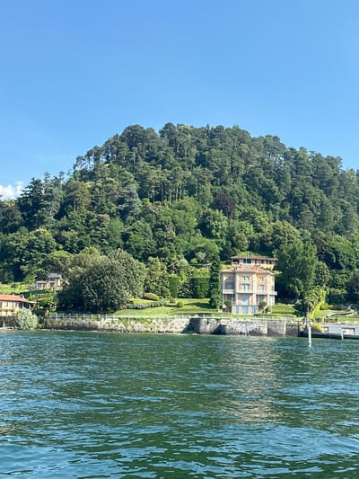 The image shows a view of Lake Como in San Giovanni, Italy, from the water. In the foreground, the deep green water of the lake is visible, with ripples and reflections of sunlight. The middle ground features a stone embankment along the shore, topped with a manicured lawn and vibrant flower beds. Several large, well-maintained trees and shrubs are scattered throughout the property.
Dominating the middle ground is a multi-story villa with a light-colored facade, featuring balconies and large windows. The villa is situated on a lush green hillside. Behind the villa, the hillside rises steeply, densely covered with a variety of green trees, creating a rich forest canopy that extends to the horizon under a clear blue sky. In the distance, a smaller building is partially visible amongst the trees. The scene is tranquil and picturesque, showcasing the natural beauty and elegant architecture of the Italian lakeside.