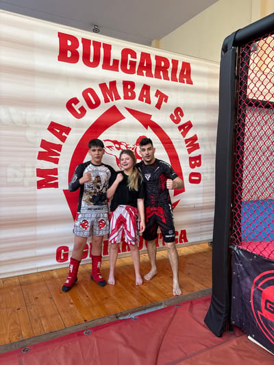Three individuals, two males and one female, stand in front of a banner that reads "BULGARIA COMBAT SAMBO" in large red letters. The banner also features a stylized red arrow. The three people are posing for a photo. The male on the left is wearing a black and white long-sleeved rashguard with graphics and grey shorts with a red and white pattern. He has red shin guards on. The female in the middle is wearing a black t-shirt and red and white striped shorts. The male on the right is wearing a black and red short-sleeved rashguard and black shorts with red accents. They are standing on a wooden floor. To the right of the individuals and banner is a portion of a black and red cage, characteristic of mixed martial arts (MMA) or combat sports. The overall scene suggests a combat sports gym or event venue. The backdrop is a plain wall. The lighting is bright and appears to be from overhead sources. The image depicts a scene likely at a combat sports facility in the city of Gorna Oryahovitsa, Bulgaria, given the prominent banner.