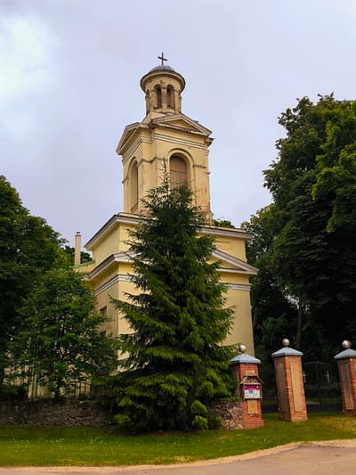 The image shows a pale yellow church with a bell tower topped with a cross. The church is surrounded by lush green trees and foliage. In the foreground, there is a brick gate with decorative caps and a notice board attached to one of the pillars. A paved road runs in front of the gate and the churchyard. The sky is overcast, suggesting a cloudy day. The scene depicts the exterior of the church, likely a historical building within the town of Vecumnieki, Latvia. There are no people visible in the image.