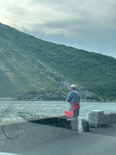 An elderly man wearing a cap, a light blue shirt, and dark shorts is standing on a stone embankment, fishing. He has a red bag slung over his right shoulder and a bright red plastic bag at his feet. His fishing rod extends into the calm, rippling water. In the background, a densely vegetated, steep green mountain rises from the shoreline, under a cloudy, overcast sky. The embankment is made of rough-hewn stone blocks and the paved road is in the foreground. The scene is likely located in Lepetane, Montenegro, given the characteristic landscape of the Bay of Kotor.