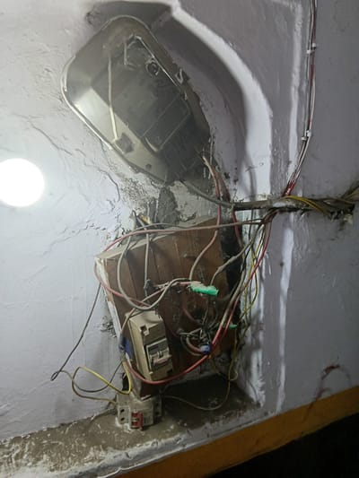 The image depicts an exposed electrical junction box mounted on a white wall, possibly a ceiling, within a building. The location is potentially in Jaipur, India, based on the image request context. The junction box is crudely constructed, seemingly from wooden material. Exposed, multi-colored electrical wires are connected to various electrical components, including switches and relays. A metal housing for some type of fixture, possibly a light, is adjacent to the junction box. The surrounding environment shows signs of disrepair with cracked paint and general grime. A light source shines brightly in the lower center, potentially an unmounted bulb. There are no people visible.