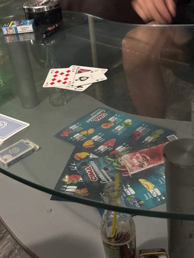 A glass-topped table in Kubrat, Bulgaria, holds various items. In the upper left, a cigarette pack and a lighter sit near an ashtray containing a lit cigarette. In the center, playing cards are spread out, two of which show the number ten of diamonds, and one of which displays an ace of spades. To the left, a deck of cards in a blue case rests next to a single opened card box labeled "PLATNIK CLASSIC". Several colorful flyers, possibly for drinks or a menu, are scattered on the table. One flyer features images of fruits like strawberries, blueberries, watermelon, and mango, with text in Cyrillic. A glass bottle with a yellow straw contains a dark red liquid, and a small QR code is visible on one of the flyers. The scene appears to be indoors, with a person's legs and hands visible in the background, suggesting a casual gathering or game.