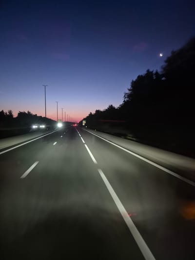 The image shows a highway scene at dusk, with the sky transitioning from a deep blue to hues of purple and pink near the horizon. The road ahead is illuminated by the headlights of oncoming vehicles and streetlights. The right side of the road is bordered by a dark line of trees. The crescent moon is visible in the upper right portion of the sky, with a faint star nearby. The highway appears to be a multi-lane road with clear white lane markings. The scene suggests a typical evening commute or travel on a highway outside of urban centers. The location is identified as the city of Schoten, Belgium.