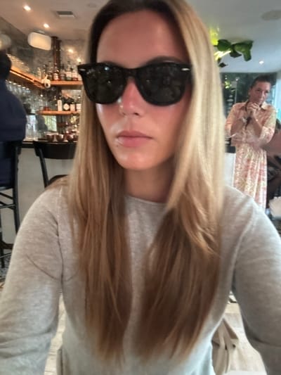 The image depicts an indoor scene within an upscale restaurant or bar located in Miami Beach, United States.
**Foreground:** A female, likely in her 20s or 30s, is positioned centrally, directly facing the camera. She has long, straight, light blonde hair. Her face is partially obscured by black Wayfarer-style Ray-Ban sunglasses. Her nose appears reddish. She is wearing a light grey long-sleeved top. Her expression is neutral.
**Background (Left):** To the left of the central subject, a well-stocked bar area is visible. Bottles of various spirits and wines are arranged on shelves against a textured, possibly stone or concrete, wall. Glasses hang inverted above the bar. A person wearing a dark blue top is partially visible, seated on a black bar stool at the counter. Recessed lighting is present on the ceiling above the bar.
**Background (Right):** To the right and behind the central subject, another female is visible, out of focus. She has dark hair, potentially pulled back, and is wearing a light-colored dress with a floral or patterned design. Her hands are clasped near her chest or chin. Beyond her, lush green foliage is discernible, suggesting an adjacent garden, patio, or outdoor area through a window, bathed in brighter light.
**Overall Environment:** The setting is characterized by a sophisticated yet relaxed ambiance, indicated by the well-appointed bar, modern lighting, and integration of natural elements. The specific establishment is not explicitly identifiable from the visual information; however, its decor and style are consistent with upscale dining and lounge venues common in areas of Miami Beach.