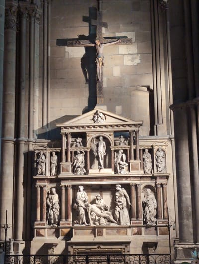 The image displays a detailed sculpture of a crucifixion scene within a church in Urvillers, France. The central element is a large wooden cross with a sculpture of Jesus Christ affixed to it. To the right of the cross, a shadow is cast onto the stone wall of the church. Below the crucifixion, a complex multi-tiered marble monument is present, adorned with numerous sculpted figures. These figures depict various religious scenes and individuals, including what appears to be a pietà on the lower tier. The monument features architectural elements like columns and pediments. The overall setting is that of a dimly lit church interior, with stone columns visible on the left and right edges of the frame, and elaborate stonework forming the backdrop. A black metal railing is visible at the bottom of the image, suggesting a roped-off area in front of the monument.