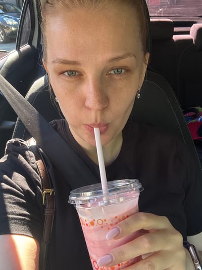 A blonde woman is in the driver's seat of a car, possibly in Fedorino, Russia, based on the request, though the visual elements do not confirm a specific city. She is drinking a pink beverage from a plastic cup with a straw. She is wearing a black t-shirt and a brown strap is visible across her chest. Sunlight is casting shadows on her face and arm. The interior of the car, including a seatbelt and headrest, is visible. The background shows other vehicles and possibly a building.