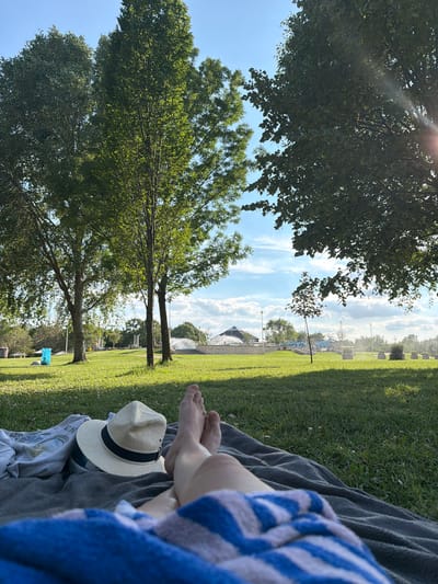 The image depicts a person relaxing on a blanket in a grassy park on a sunny day. In the foreground, the person's legs and feet are visible, covered partially by a blue and white striped towel. A straw hat with a black band rests on a gray blanket next to a crumpled white garment.

The background shows a well-maintained park with lush green grass, several trees with dense foliage, and a bright blue sky with scattered white clouds. In the mid-ground, there are structures that appear to be part of a public facility, possibly a water park or a fountain area, with water spraying in the distance. Lampposts and low-lying stone planters are also scattered throughout the park. A bright blue plastic bag or container is visible on the grass to the left of the frame. The overall impression is one of leisure and outdoor recreation in an urban park setting.

While the image provides a visual representation of a park scene, it does not contain any specific information or landmarks that would allow for definitive identification of the location within the city of Kiskunmajsa, Hungary. The scene is generic to many parks found in temperate climates during warmer months.