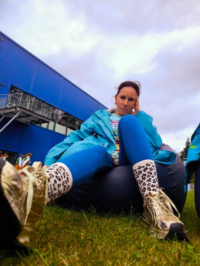 A low-angle outdoor photograph captures a woman seated on a dark bean bag on a vibrant green lawn. She is wearing a light blue raincoat over a lighter blue t-shirt, royal blue leggings, cream-colored socks with a leopard print, and metallic gold athletic shoes with matching laces. Her right hand is positioned near her temple, and she looks directly at the camera with a focused expression. A red earring is visible on her left ear.
In the background, a large, multi-paneled building with a distinct blue exterior dominates the left side of the frame. This structure is identified as the IKEA store located at Biķeru iela 2 in Rīga, Latvia. The building features an external metal staircase with railings leading to an upper level with large windows. To the far left, near the base of the building, several individuals are visible, dressed in traditional Latvian folk costumes consisting of green vests, white blouses, patterned skirts, and floral wreaths on their heads, suggesting a cultural event or gathering. The sky above is overcast and filled with dense clouds. A cluster of trees is visible in the background on the right side of the image.