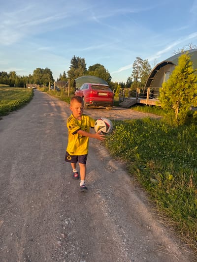 A young boy walks down a dirt road in Zamosh'e (Zaklinskoe S/P), Russia, holding a soccer ball. He is wearing a yellow jersey with blue shorts and dark blue slip-on shoes with a red star design. The road is bordered by green grass and trees on either side. In the background, there are several glamping tents and a red car. The sky is blue with some wispy clouds, and the sun is setting, casting long shadows.