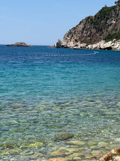 The image depicts a serene coastal scene at Perazića Do, Montenegro, with the camera oriented vertically, making the water appear to be flowing from left to right across the frame. The left side of the image features a rugged, rocky cliff face covered in green vegetation, sloping down towards the sea. A clear blue sky is visible above the cliff.
The water itself is the dominant subject, transitioning from a deep blue hue near the shore to a more transparent, turquoise color further out. The transparency of the water allows the rocky seabed and scattered stones beneath the surface to be clearly visible, creating a mesmerizing, rippled effect.
Along the boundary between the darker and lighter blue water, a white, dashed line of buoys stretches across the frame, likely marking a swimming area or a boundary for water activities. Several dark, small objects, possibly swimmers or small boats, are visible in the water near the buoys.
The overall impression is one of a calm and clear day, likely mid-morning or early afternoon given the bright sunlight reflecting off the water and illuminating the seabed. The location is identifiable by the distinctive rocky cliffs and the characteristic clear, blue waters of the Montenegrin coast. There is no visible text in the image.
