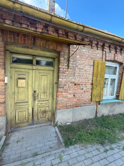 This image shows the exterior of a weathered brick building in Sobolewo, Poland, on a clear day under a bright blue sky. The building features a vintage, pale yellow double door with intricate carvings, a small, weathered mailbox attached to the left door, and a decorative transom window above. To the right of the door is a window with a light blue frame and a distressed yellow wooden shutter. The brickwork of the building appears aged, with some signs of wear and mortar degradation. The roofline has a yellow gutter. The ground in front of the building is paved with gray interlocking bricks, with patches of green grass and dry leaves growing in between and around the paving stones.