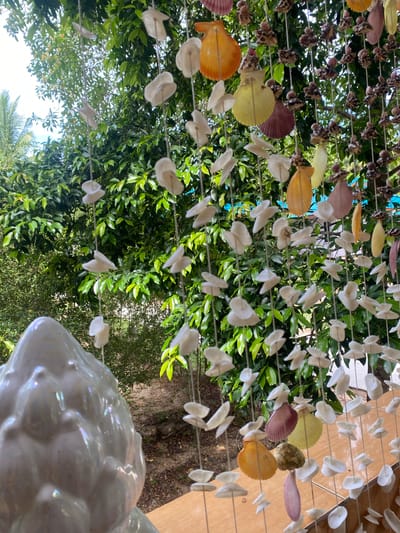 The image depicts a close-up, upward-facing view of a seashell wind chime hanging outdoors in Baan Nai Suan, Thailand. The wind chime is constructed with multiple strands of rope, each adorned with various seashells in colors like white, yellow, orange, brown, and purple. Some shells are small and singular, while others are larger and appear to be scallop shells. The background consists of lush green foliage, primarily tree leaves, filling the frame. In the bottom right corner, a portion of a white, possibly ceramic, object is visible, exhibiting a glossy, reflective surface. The overall lighting appears to be natural daylight, casting shadows through the leaves and highlighting the sheen of the shells. The image has been rotated.